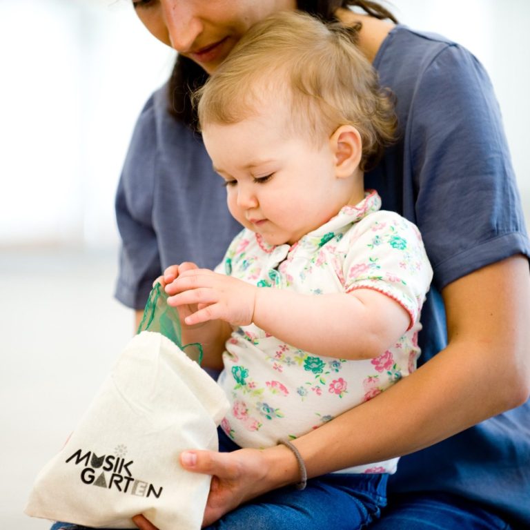 Ein Baby entdeckt die Musikgarten-Materialien. Ein Baby sitzt auf dem Schoß der Mama und entdeckt sein Musikgarten Säckchen. Es zieht ein Tuch heraus.